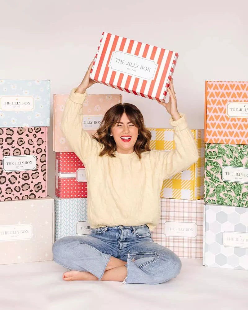Jillian Harris sitting cross-legged holding a Jilly Box above her head with various Jilly Boxes in the background